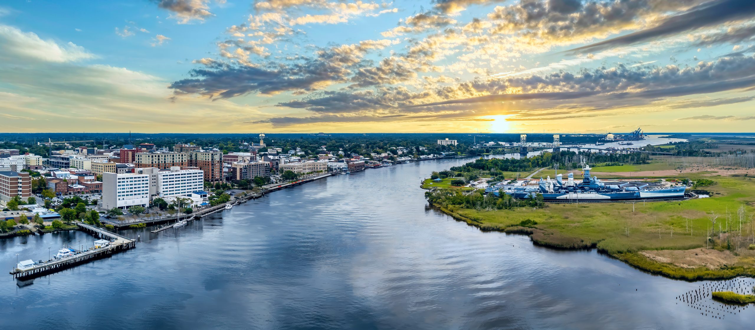 Aerial cityscape along a wide river at sunset, with buildings lining the waterfront and a distant harbor on the right.
