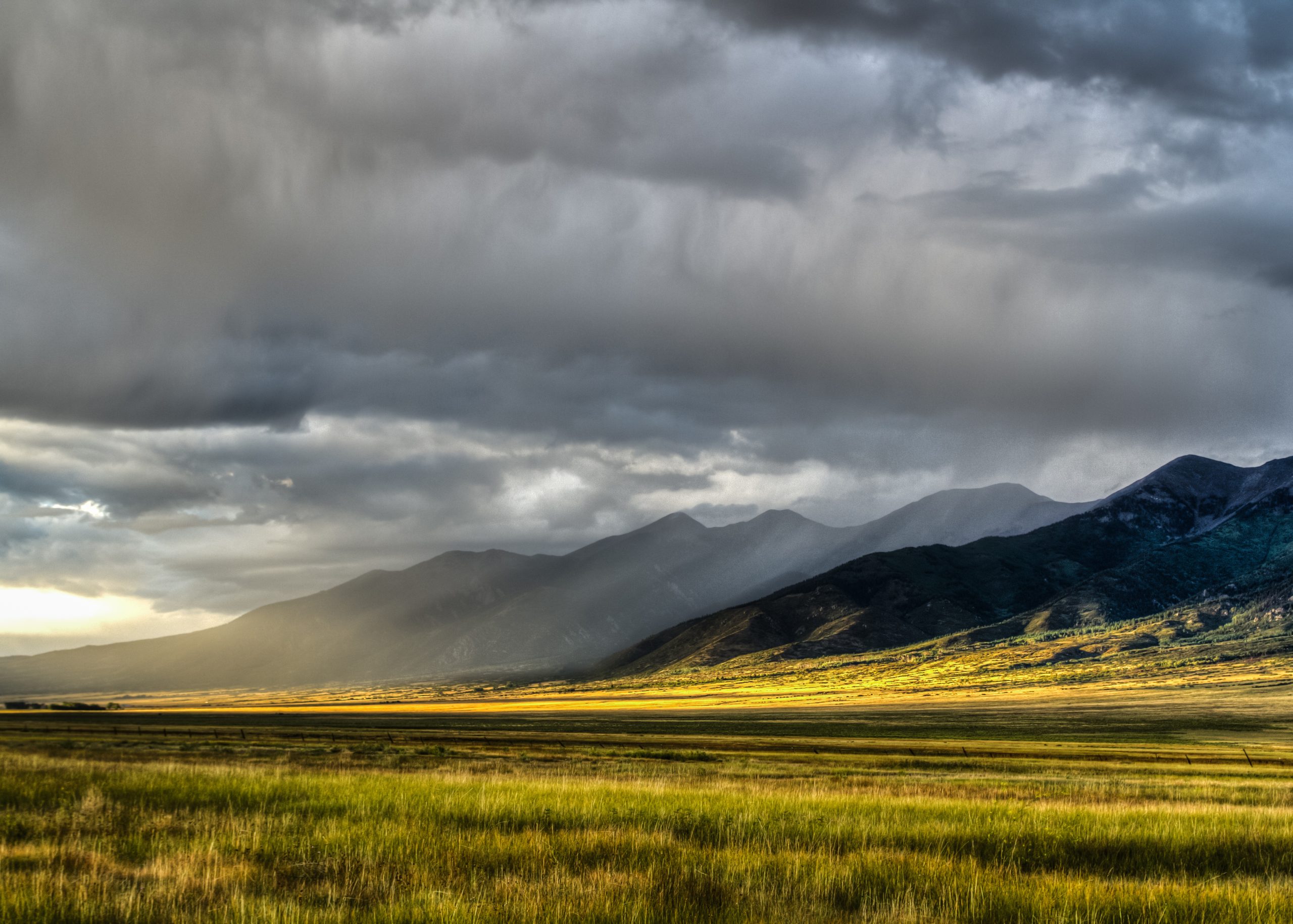 Wide valley with sunlit grass in the foreground and misty mountains under a cloudy sky.