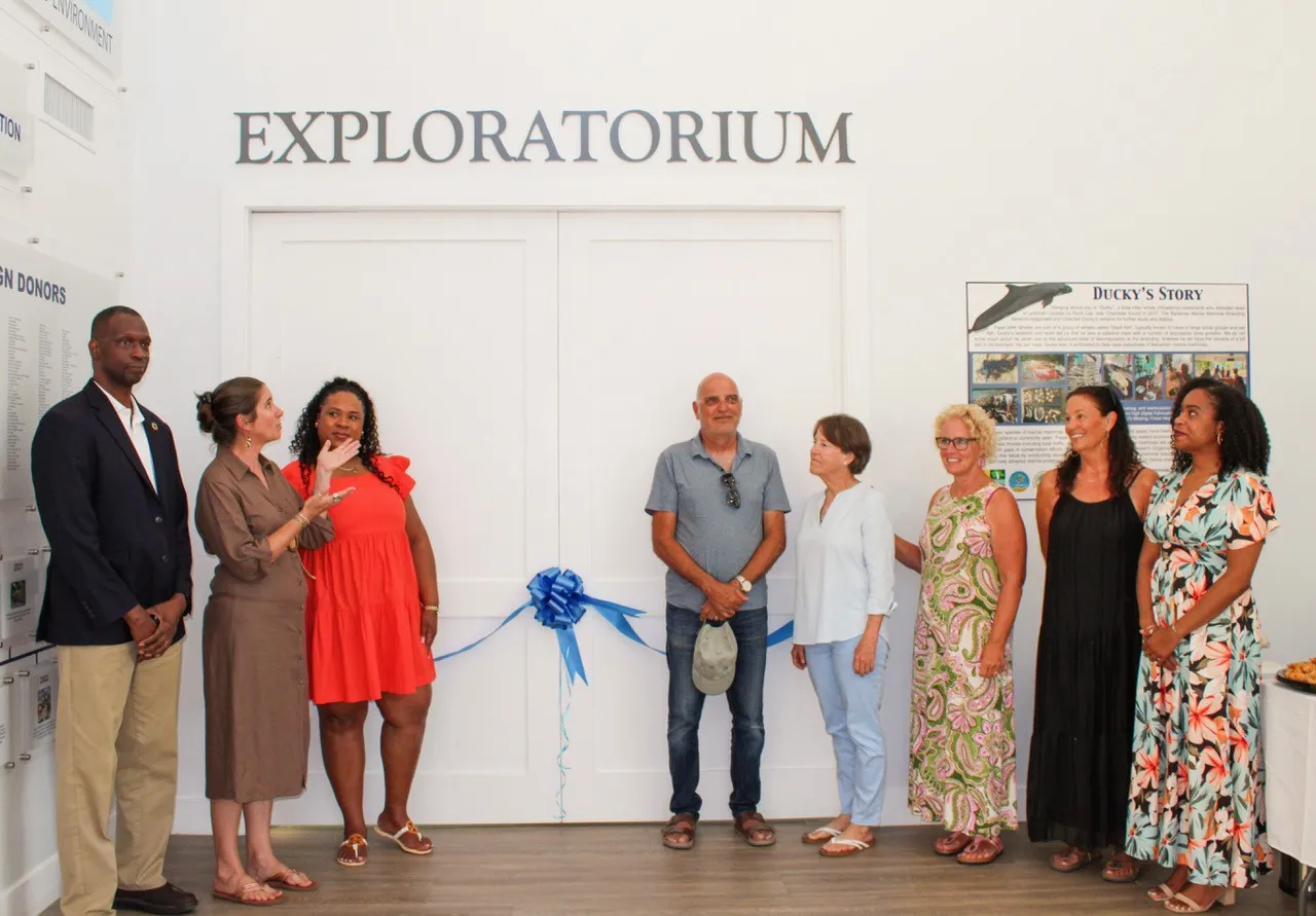 Group of diverse adults standing in front of a white wall with 'EXPLORATORIUM' sign, blue ribbon for a ribbon-cutting ceremony snapping a moment of opening.