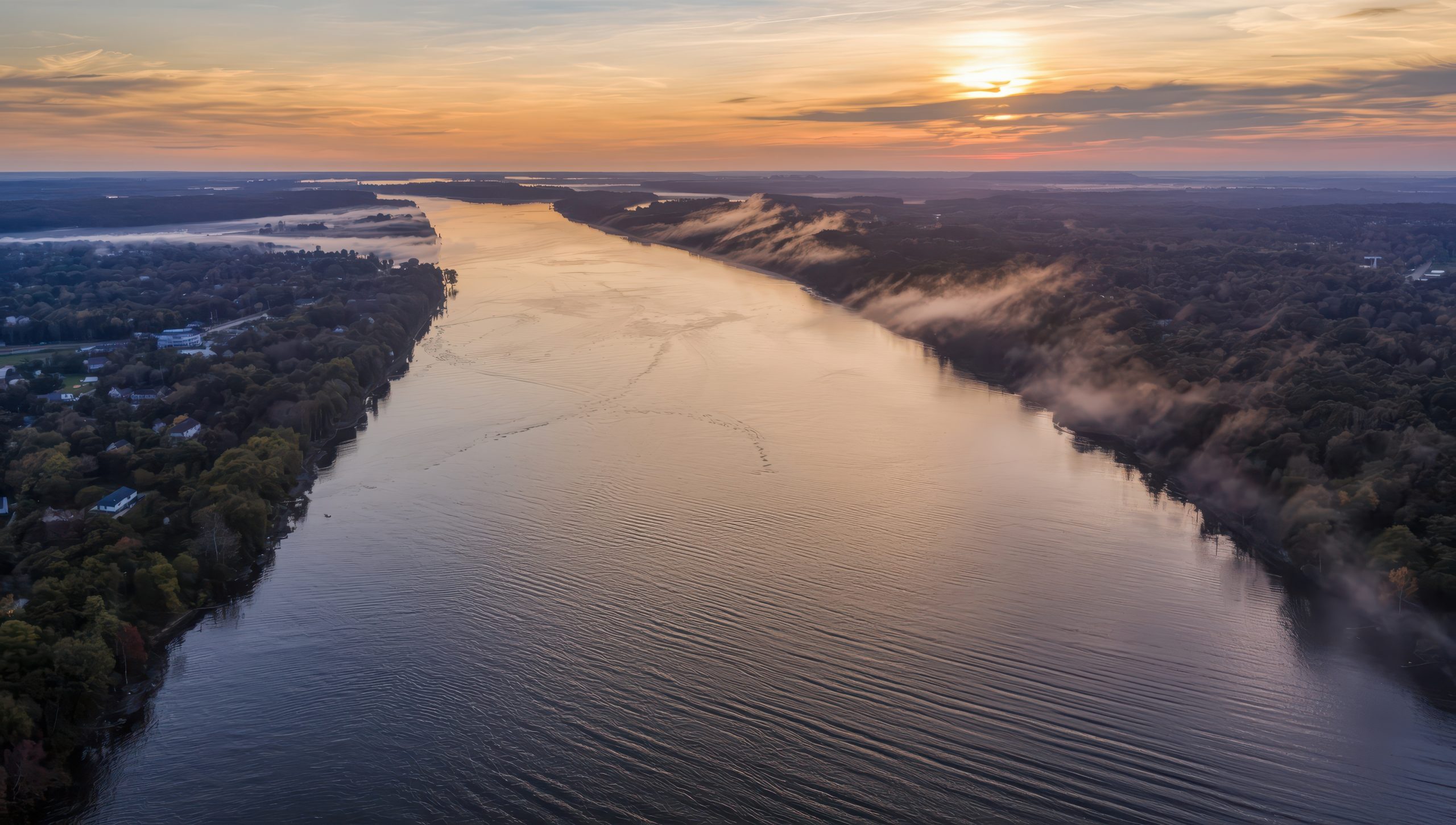 Aerial view of a wide river winding between forested banks at sunset, with light mist along the shore.